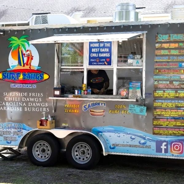 Surf Dawgs Grill food truck with a server inside, menu boards, and branding visible.