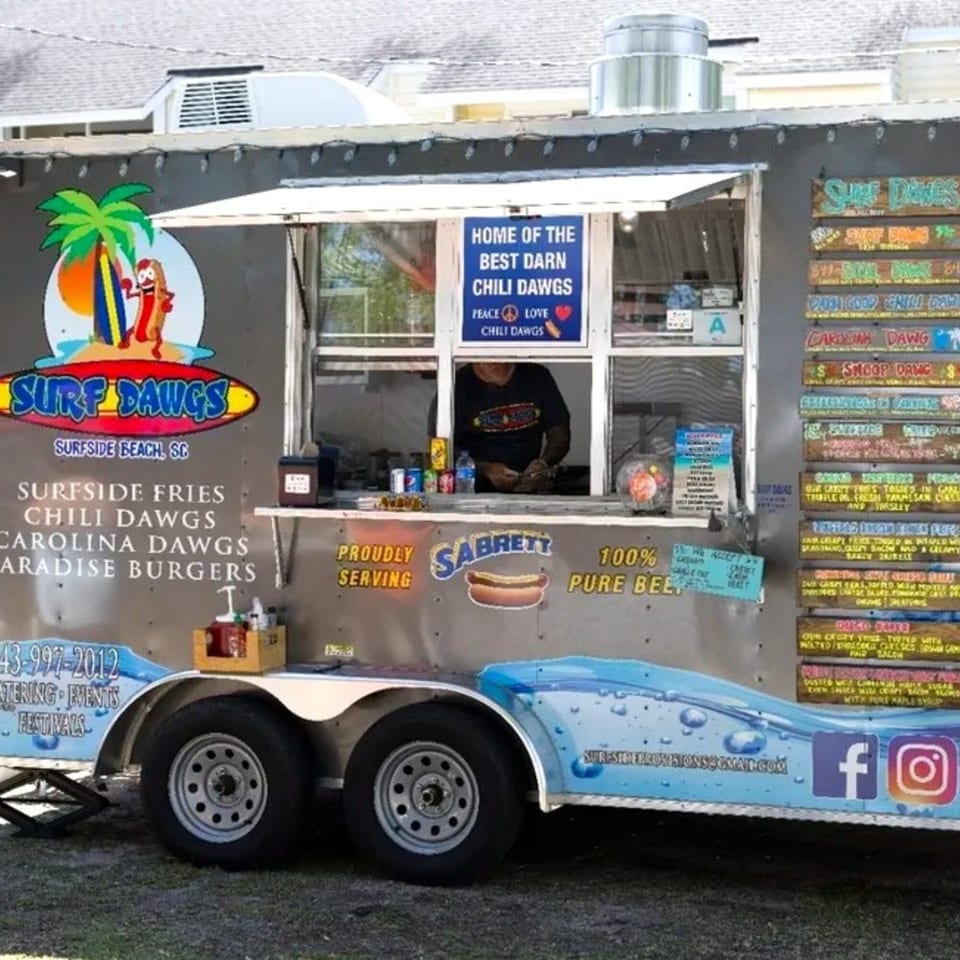 Surf Dawgs Grill food truck with a server inside, menu boards, and branding visible.