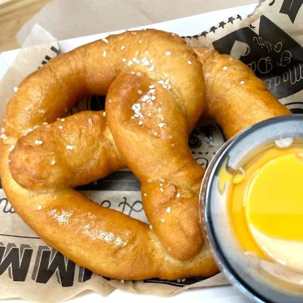 A golden-brown, salted pretzel with a side of yellow cheese dip on branded paper.
