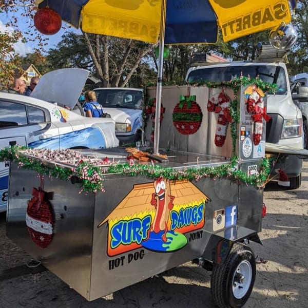 Surf Dawgs Grill hot dog cart decorated with Christmas garland and ornaments, parked outdoors.
