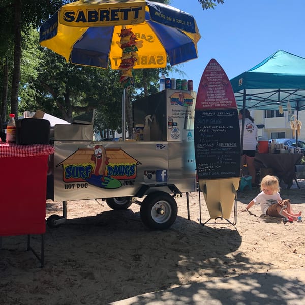A Surf Dawgs Grill hot dog cart with a yellow umbrella, menu board, and a child playing in sand.