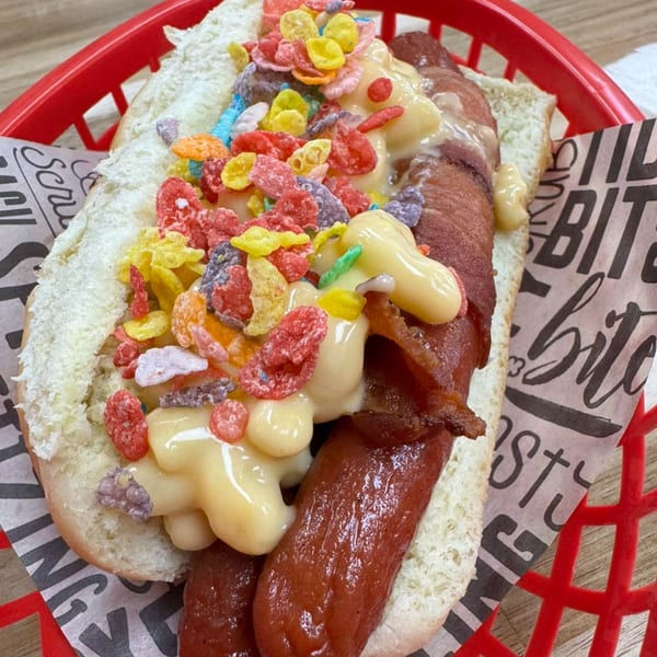 A Surf Dawgs Grill hot dog with bacon, mac and cheese, and colorful cereal in a red basket.